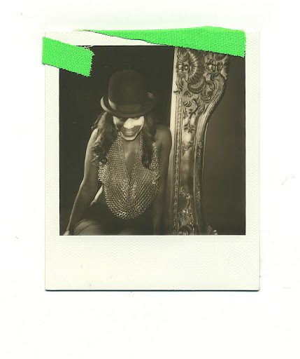Polaroid photo of woman in chainmail top and bowler hat sitting on ornate chair with slight smile, vintage style, good-times feeling.