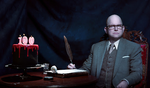 Adult male at table with cake, quill pen writing on parchment, in front of a dark draped background, wearing glasses and suit.