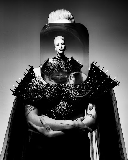 High contrast portrait of a non-binary person with short, light hair wearing dark, spiked epaulettes and cape in an indoor studio.