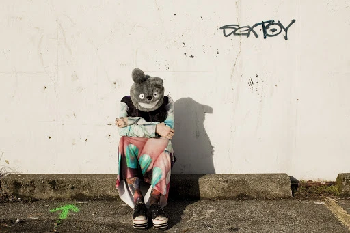 Person wearing a bear mask sits outside against a white wall with graffiti, wearing colorful clothing and black platform shoes.
