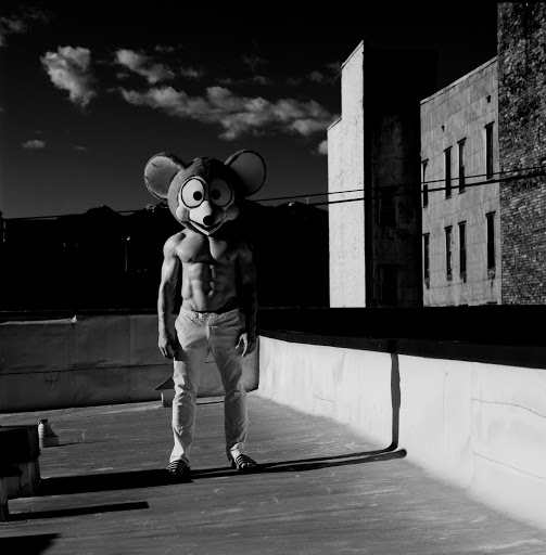 Person in a cartoon mouse costume with exposed abs standing on a rooftop with city buildings and cloudy sky in the background in black and white.