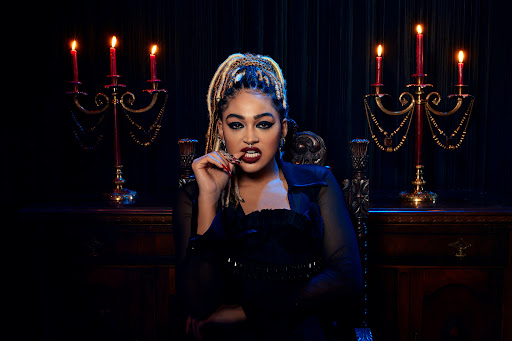 Woman in gothic style dress sits in ornate chair with candelabras and dark background, biting fingers, wearing jewelry, heavy makeup, braids