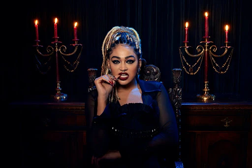 Woman in gothic style dress sits in ornate chair with candelabras and dark background, biting fingers, wearing jewelry, heavy makeup, braids