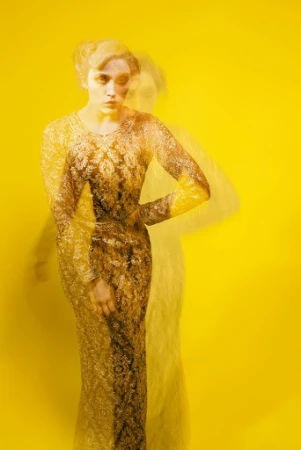 A woman in a patterned gold dress captured with multiple exposure movement against a vibrant yellow background.