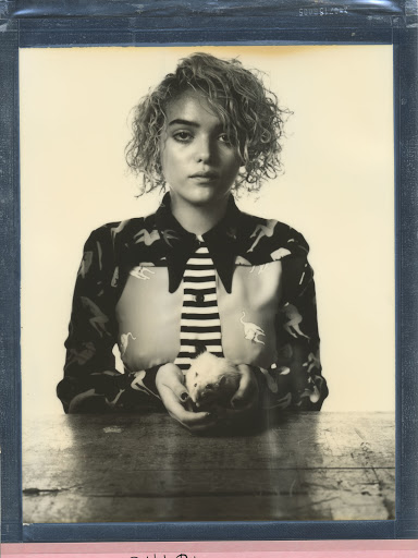 Portrait of a young woman with short curly blonde hair wearing a patterned shirt and tie, holding an apple on a wooden table.
