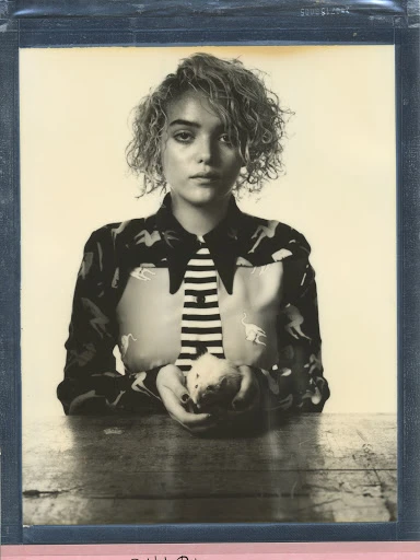 Portrait of a young woman with short curly blonde hair wearing a patterned shirt and tie, holding an apple on a wooden table.