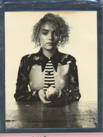 Portrait of a young woman with short curly blonde hair wearing a patterned shirt and tie, holding an apple on a wooden table.