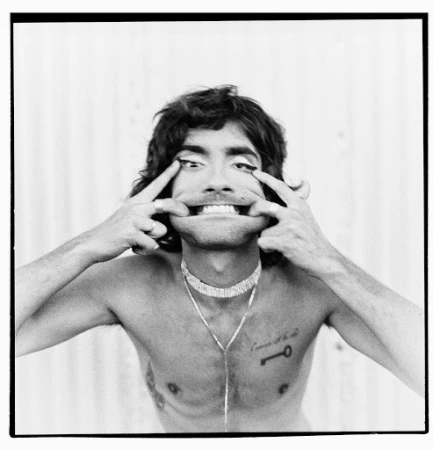 Monochrome portrait of a shirtless man pulling his mouth wide with his fingers, wearing necklaces against a ribbed backdrop.