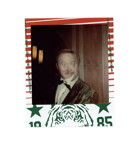A person in a tuxedo is posing for an indoor snapshot against a dark wood paneled wall with a tiger mascot and school name border.