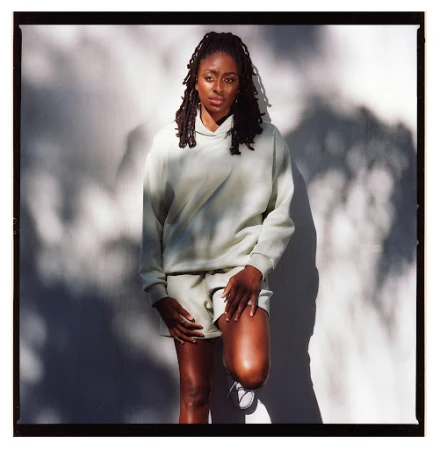 Black woman with dreadlocks wearing light green hoodie and shorts standing against a wall with harsh leaf shadows.