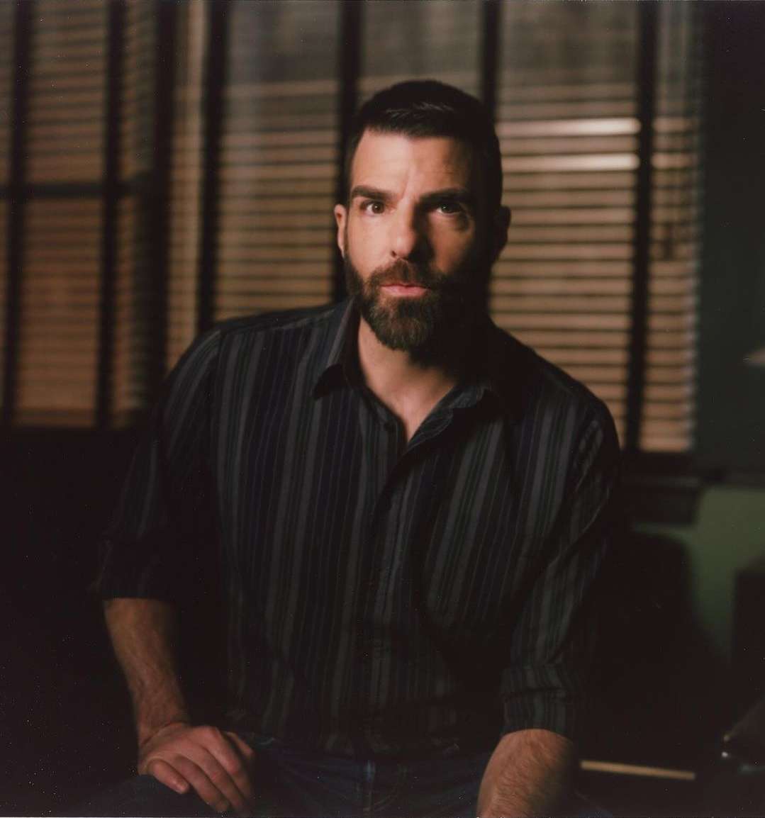 Portrait of Zachary Quinto with a short beard wearing a dark striped button-down shirt indoors against window blinds.