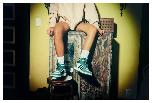 Person sits on vintage cabinet wearing green and white high-top sneakers, white socks, and sweatshirt in a warm-toned indoor setting.