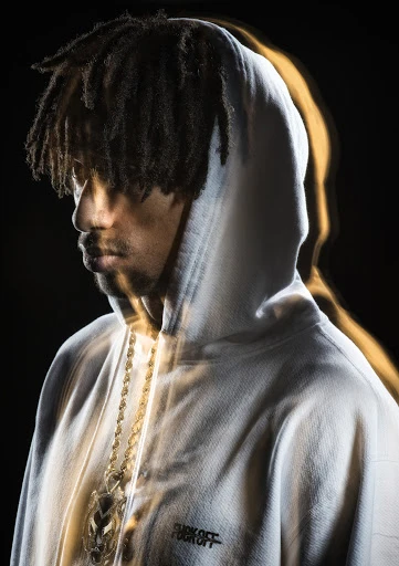 Adult man in a hoodie with dreadlocks, a gold chain, and dramatic lighting against a dark background. Fashion photography.