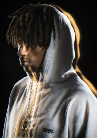Adult man in a hoodie with dreadlocks, a gold chain, and dramatic lighting against a dark background. Fashion photography.