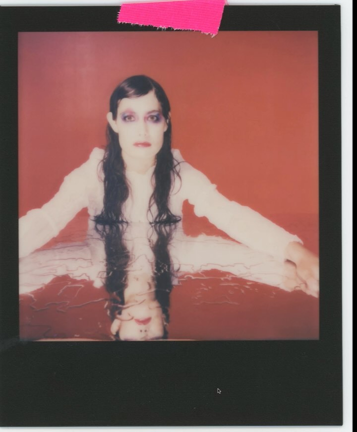 Eye-level portrait of a woman with long, dark hair lying in reddish water, with an inverted reflection, against a solid red background.