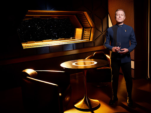 Man in blue Starfleet uniform holds a champagne glass in a lounge with a window to space, with a table and chair in the foreground.