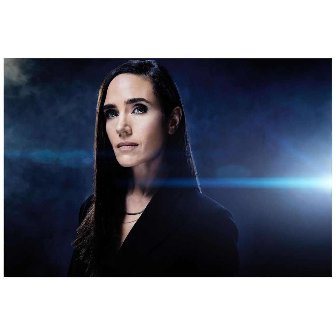 Jennifer Connelly portrait. Brunette woman in dark jacket gazes at the camera against a smokey blue and black background with lens flare.