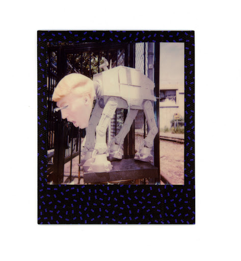 Surreal image of a human head atop an AT-AT Walker-like structure, set against an outdoor background with a chain-link fence.