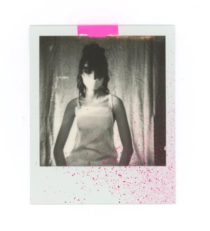 Polaroid photograph of a woman wearing a light strap top against a textured fabric curtain with pink paint splatter.