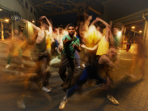 Blurred motion long-exposure photograph of dancers in an outdoor urban parking area at night with a man in green at center focus.