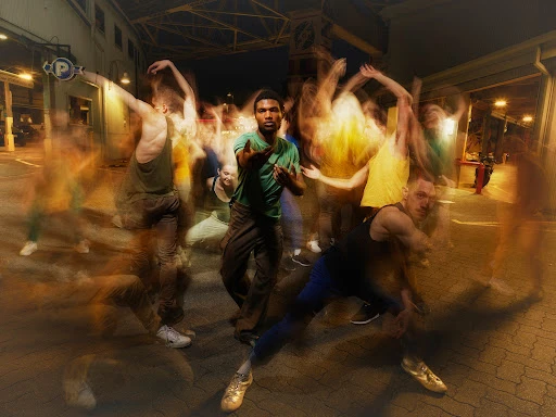Blurred motion long-exposure photograph of dancers in an outdoor urban parking area at night with a man in green at center focus.