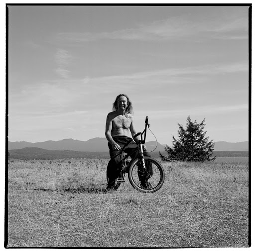 Monochrome portrait of a shirtless man with long hair on a BMX-style bicycle in a grassy field with distant mountains under a bright sky.