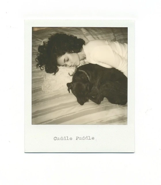 A woman with curly hair sleeps on a striped bed with a black dog curled up next to her in a warm-toned indoor shot.