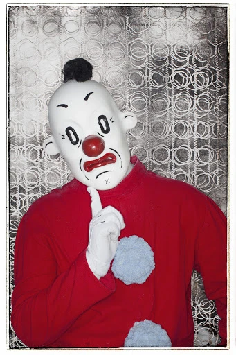 Person in clown mask and red suit points up in front of a patterned background, creating an unusual and intriguing portrait. 
