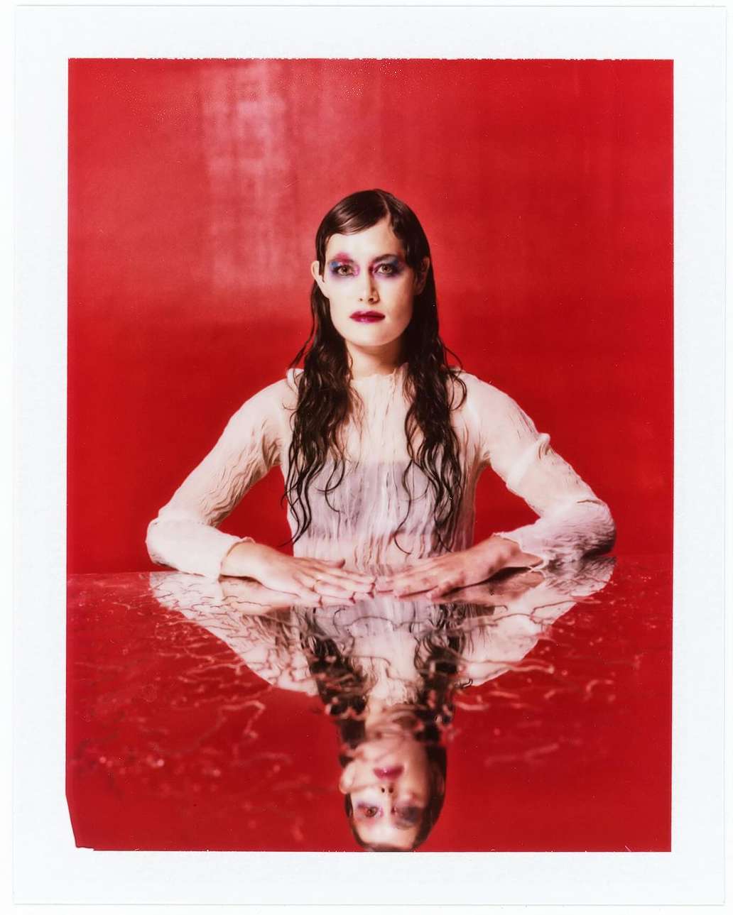 Portrait of a fair-skinned young adult woman with wet brunette hair, sitting at a reflective surface in front of a vivid red backdrop.