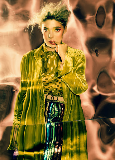 A woman in colorful fashion poses in front of a reflective background. She wears a chartreuse velvet jacket and a yellow-and-black patterned shirt.
