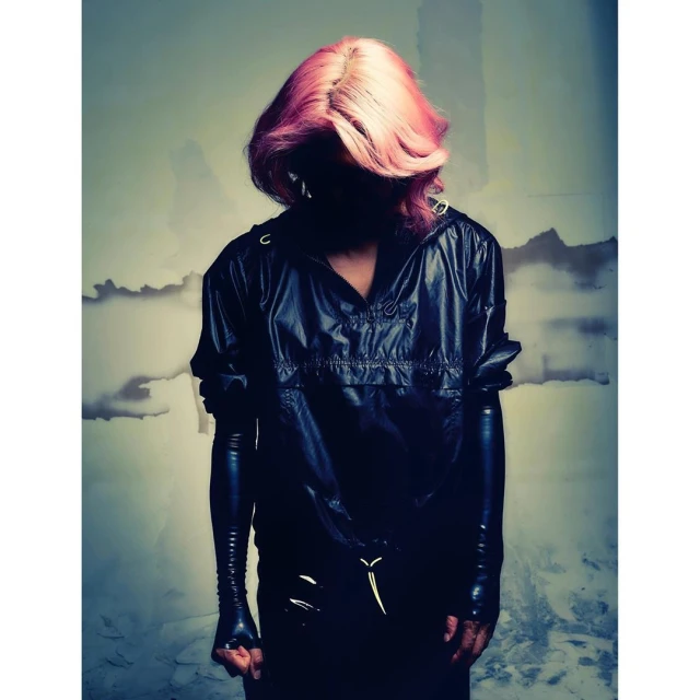 Person in dark clothing and pink hair poses against a textured background, showcasing fashion and style in a studio setting.