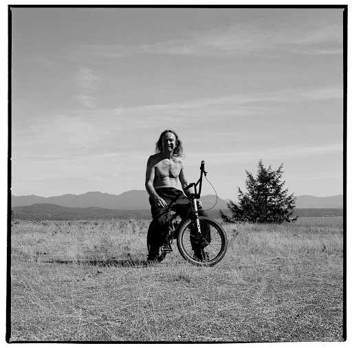 Man with long hair sitting shirtless on a BMX bicycle in an open grassy field with mountains in the background