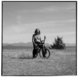 Man with long hair sitting shirtless on a BMX bicycle in an open grassy field with mountains in the background