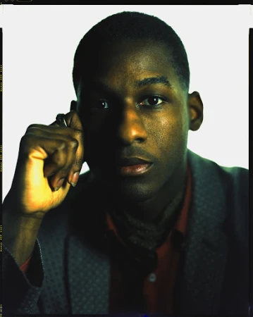 Close-up portrait of a Black man with a serious expression, wearing a patterned grey blazer and red shirt, shot by Brendan Meadows.