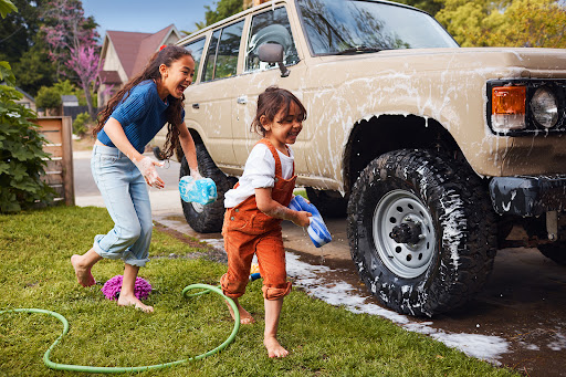 Two young girls washing a beige SUV on a grassy lawn with a green hose, enjoying a sunny day outdoors with residential houses visible.