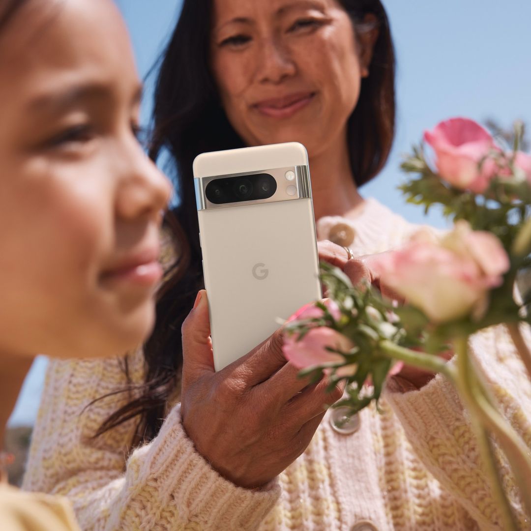 Close-up of a white Pixel phone being held by an adult, with a child and an adult woman in the background. Pink flowers are visible.