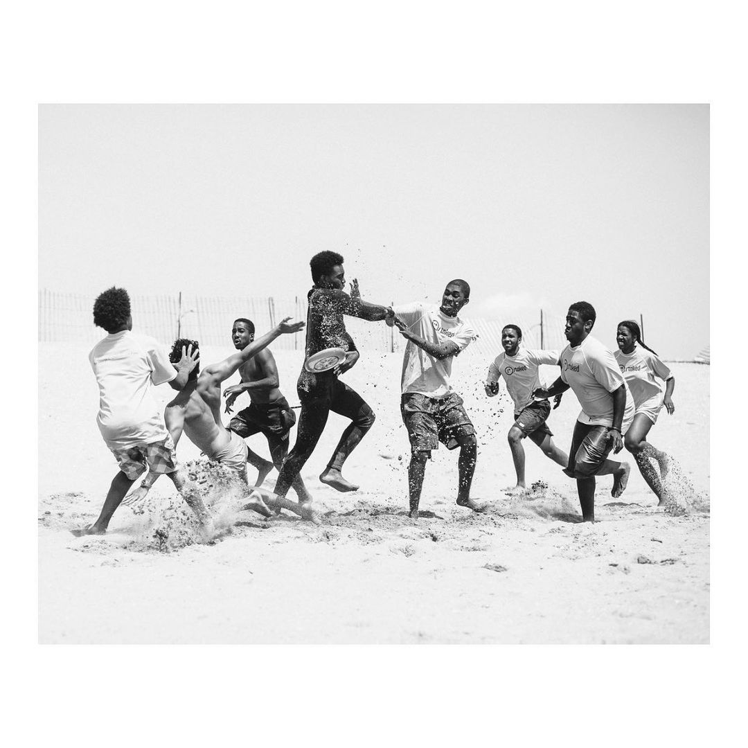 Black and white photo of a group of young adult males playing with a frisbee on a sandy beach, captured in an eye-level, wide shot.