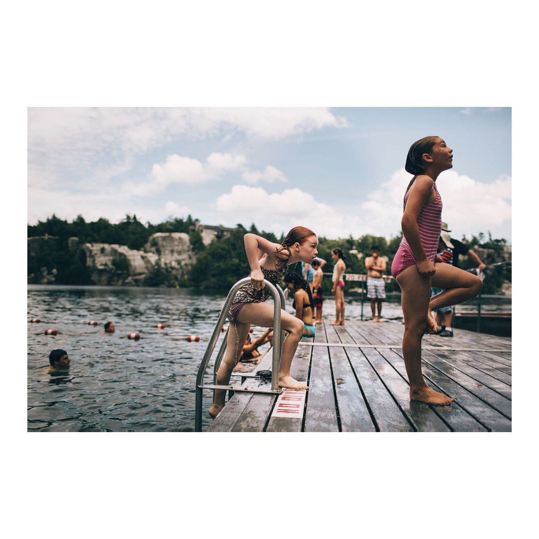 Two girls in swimsuits on a wooden dock by a quarry lake, one climbing a ladder out of the water, the other standing on the dock.