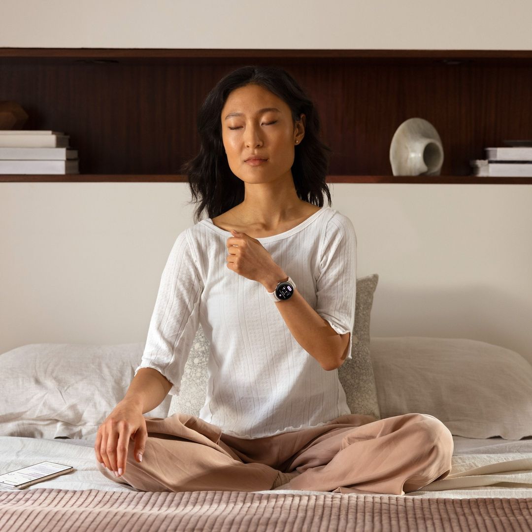 Asian woman meditating in bed with smartwatch on wrist, eyes closed, wearing casual clothes, serene expression, cozy indoor setting.