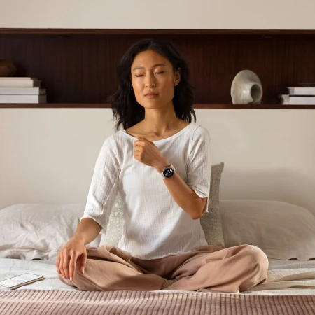 Asian woman meditating in bed with smartwatch on wrist, eyes closed, wearing casual clothes, serene expression, cozy indoor setting.