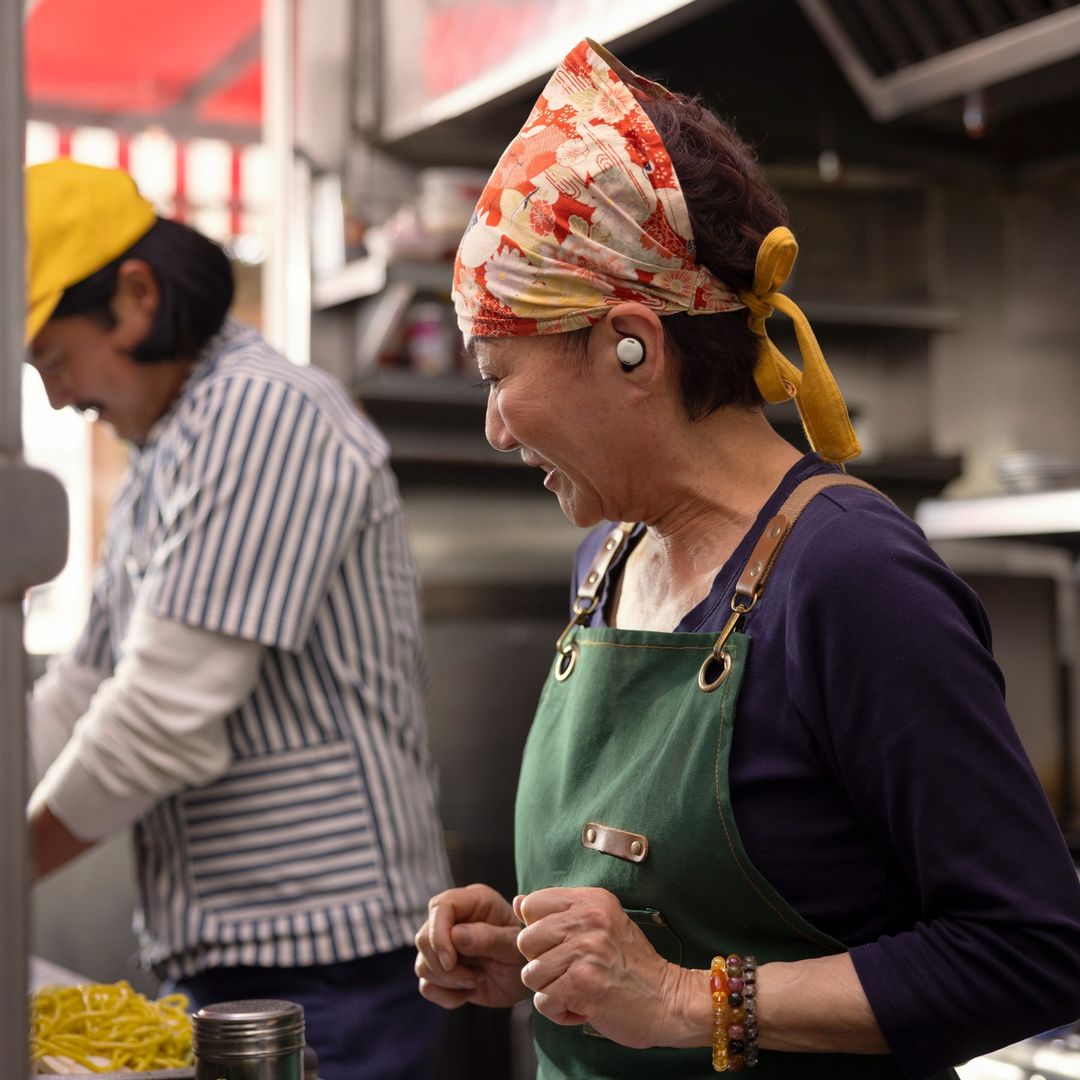 Two cooks in a restaurant kitchen: one laughing in foreground with a headscarf and apron, another preparing food in background.