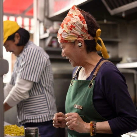 Two cooks in a restaurant kitchen: one laughing in foreground with a headscarf and apron, another preparing food in background.