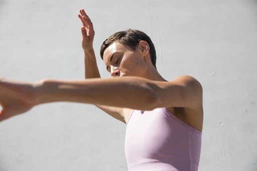 Woman with short dark hair in a pink sleeveless top posing with arms outstretched against a plain light wall