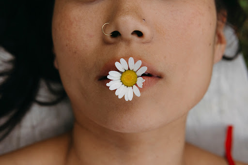 Close-up portrait of a woman with a daisy in her mouth, focusing on her face, nose ring, and shoulders, with a blurred background.