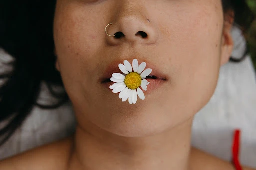 Close-up portrait of a woman with a daisy in her mouth, focusing on her face, nose ring, and shoulders, with a blurred background.