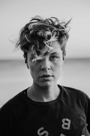 Close-up monochrome portrait of a person with short textured hair wearing a dark t-shirt with partial lettering against an outdoor horizon background.