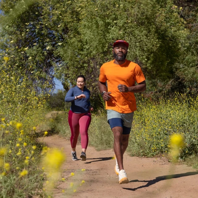 A couple runs on a dirt path surrounded by yellow flowers and green trees, with a man in the foreground and a woman behind him.