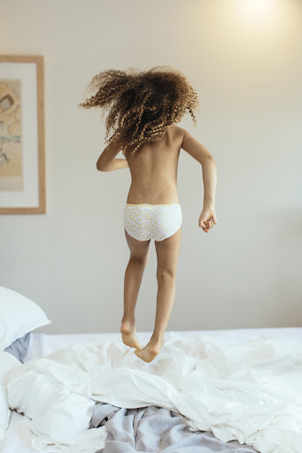 A young child with curly hair leaps on a white bed in a brightly lit room, captured from the rear with focus on the child's motion.