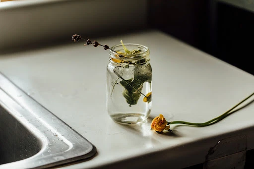 Floral arrangement featuring yellow blossoms and sprigs of lavender in a clear mason jar filled with water sitting on a white countertop near a sink.