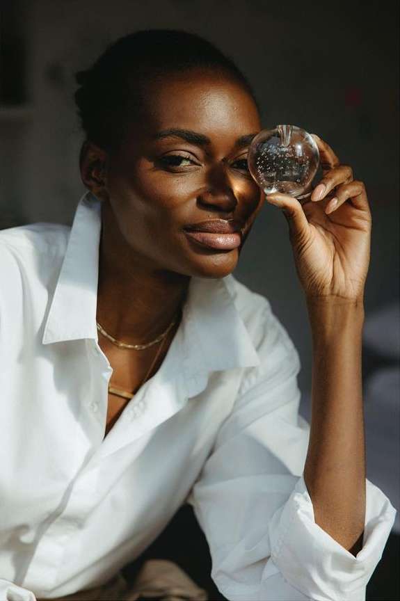 Portrait of an African American woman in a white collared shirt holding a glass orb in front of her eye, showcasing a bright indoor scene.